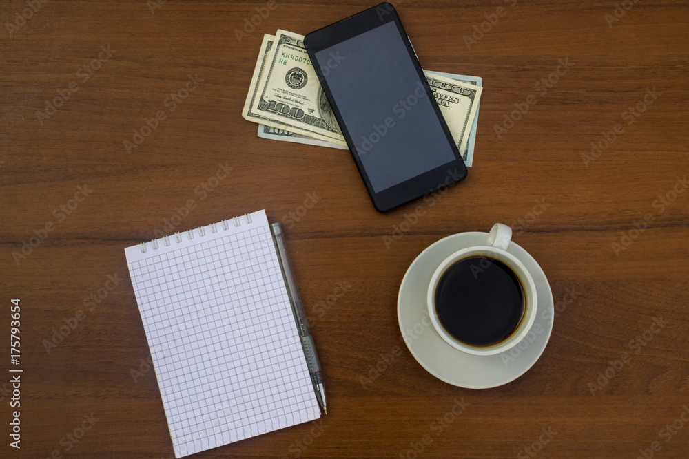 Cup of coffee, smartphone, dollars, notepad and pen on wooden desk Cup of coffee, smartphone, dollars, notepad and pen on wooden desk