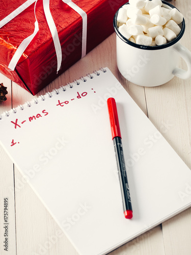 Christmas to-do list. Xmas gifts shopping planning. Make shopping or to-do list for Christmas. Notebook, mug hot chocolate with marshmallows and New Year's gift on white wooden background.