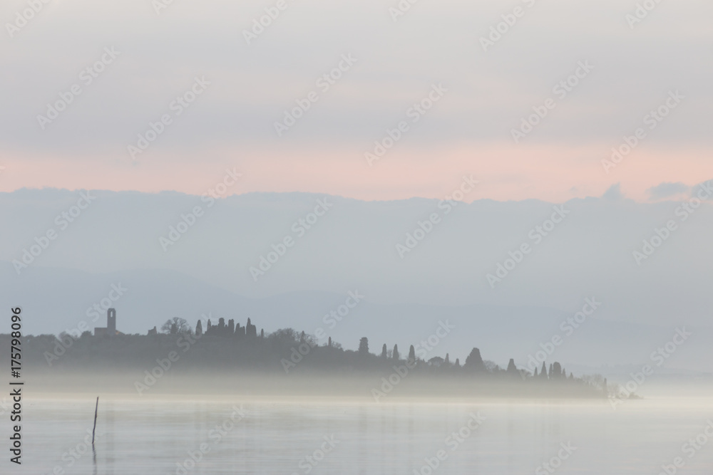 A lake at dusk, with beautiful, warm tones in the sky and water and an island with buildings in the middle of mist