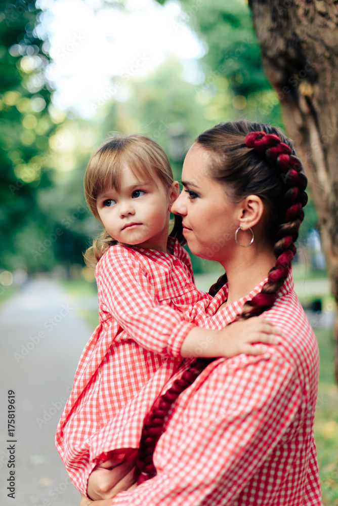 Obraz premium young happy family with little beautiful baby with blue eyes walking in summer park at sunset. Mom and little daughter in identical pink dresses