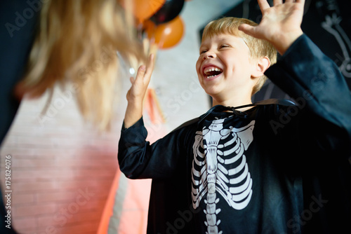 Waist-up portrait of laughing little boy wearing skeleton costume enjoying Halloween home party, blurred background