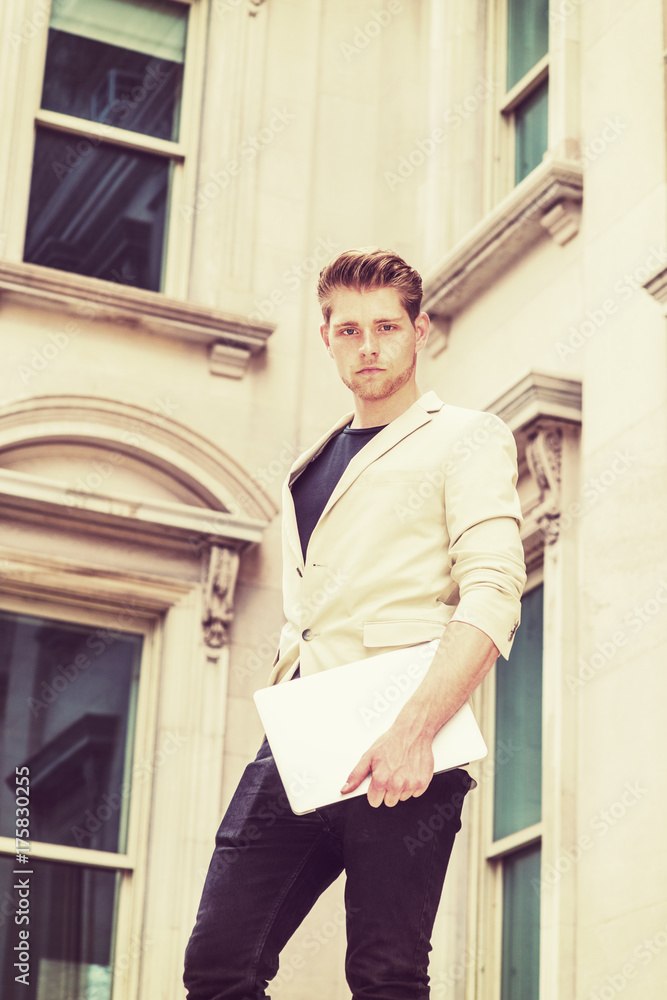 Young American Man with little beard studying, working in New York, wearing beige blazer, black pants, holding laptop computer, standing outside vintage office building on campus, looking at you.. Young American Man with little beard studying, working in New York, wearing beige blazer, black pants, holding laptop computer, standing outside vintage office building on campus, looking at you..