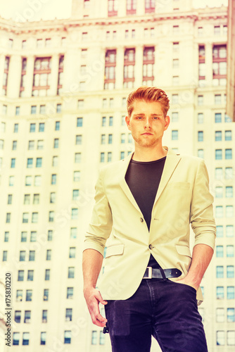 Young American Businessman with little beard traveling in New York, wearing beige blazer, black undershirt, pants, standing on street by vintage high business building, thinking. filtered effect.