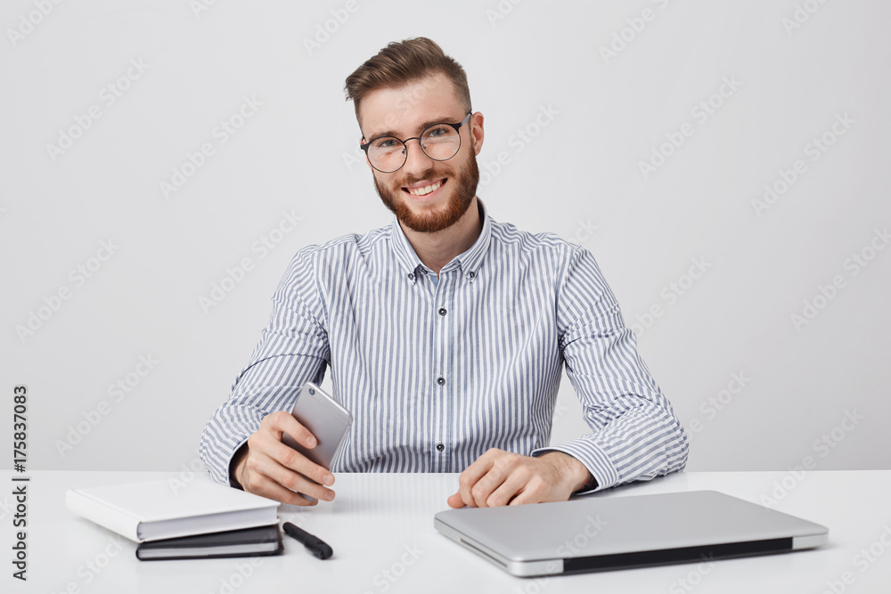 Candid shot of successful male with thick beard, uses modern technology for work, reads sms on cell phone, being glad, messages online uses free internet connection. People, business concept Candid shot of successful male with thick beard, uses modern technology for work, reads sms on cell phone, being glad, messages online uses free internet connection. People, business concept
