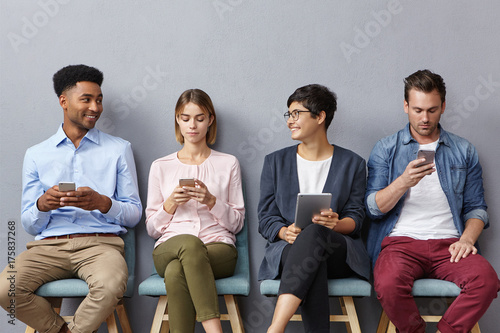 Diverse group of young people have lively conversation as sit in queue, use modern gadgets for different goals: communication, entertainement, searching information, surfing social networks.