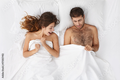 Picture of adult European bearded man and excited woman lying in bed and peeping under white blanket. Couple going to have sex in white bedrrom or hotel room. Love and relationships problems photo