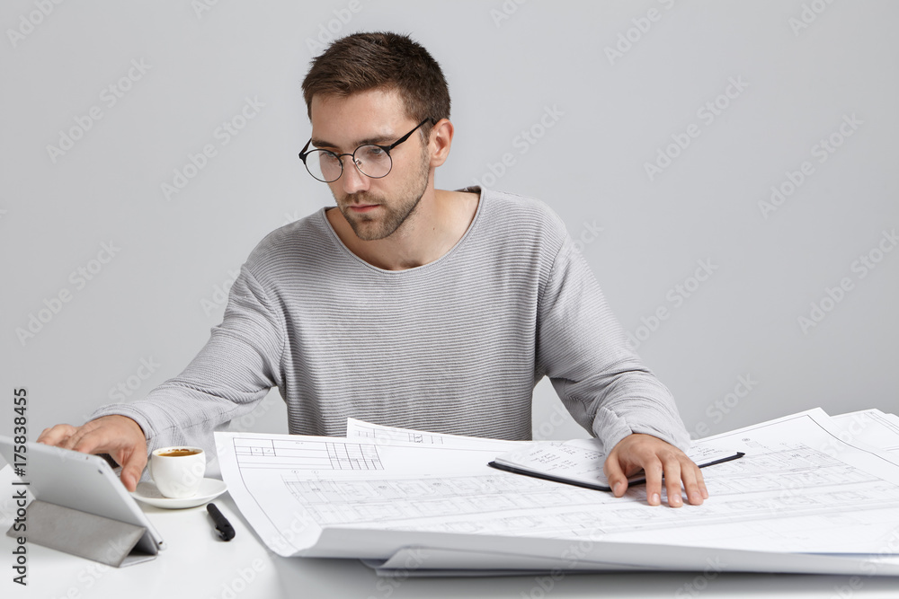 Confident male worker looks attentively into tablet computer, work at construction project, drinks espresso or capuccino. Talented young designer works alone at workplace, has serious expression Confident male worker looks attentively into tablet computer, work at construction project, drinks espresso or capuccino. Talented young designer works alone at workplace, has serious expression