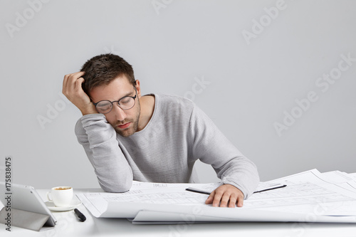 Tired young attractive man sleeps at work place, has much work, being fatigue and exhausted, isolated over white studio background. Sleepy male engineer has deadline, busy all day, has sleppless night