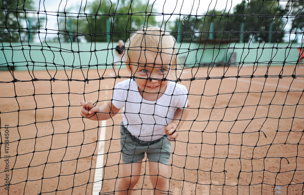 Portrait of the smiling child through a tennis net. Little cute girl in a white T-shirt at tennis court.