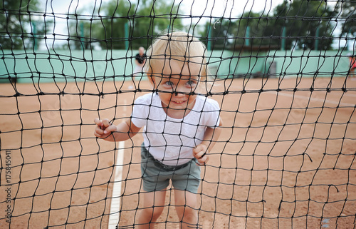 Portrait of the smiling child through a tennis net. Little cute girl in a white T-shirt at tennis court.