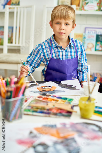 Portrait of blonde little boy looking at camera while painting Halloween pictures sitting at table in art class