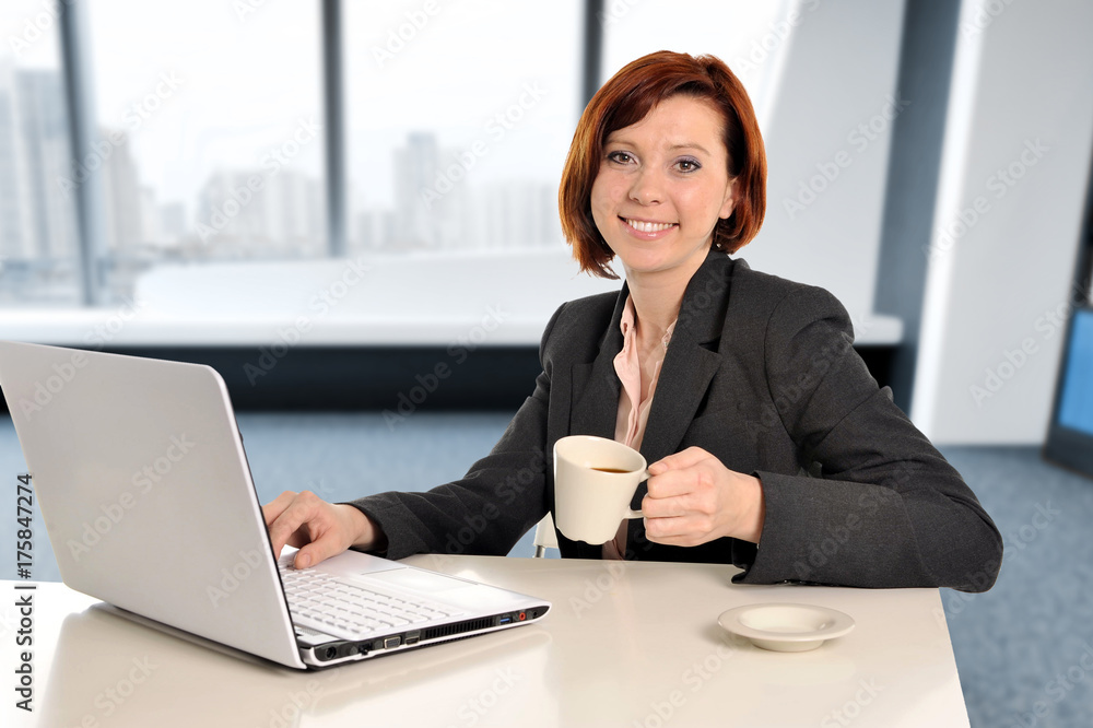 business woman with red hair at work smiling on laptop computer desk and drinking coffee business woman with red hair at work smiling on laptop computer desk and drinking coffee