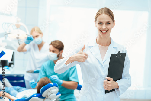 A nurse posing against a background of dentists who treat a girl's teeth. It shows thumbs up