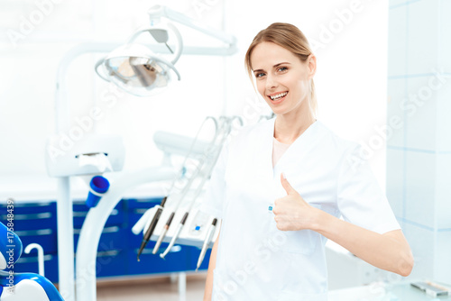 Female dentist posing against a background of dental equipment in a dental clinic. She show thumbs up
