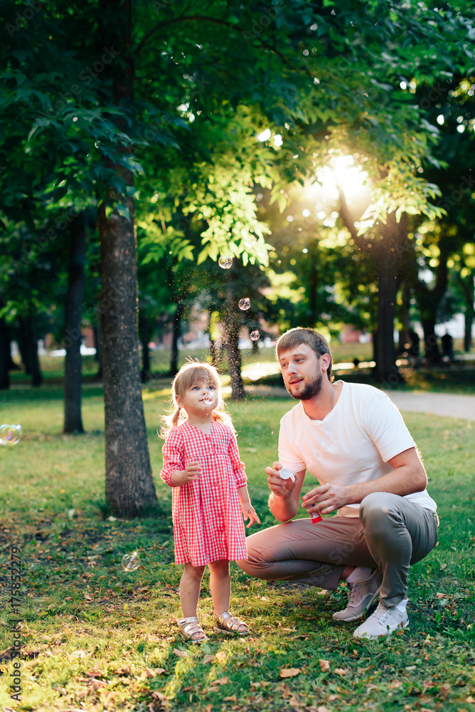 Fototapeta premium happy father with a small daughter at sunset blowing soap bubbles in a summer park outdoors