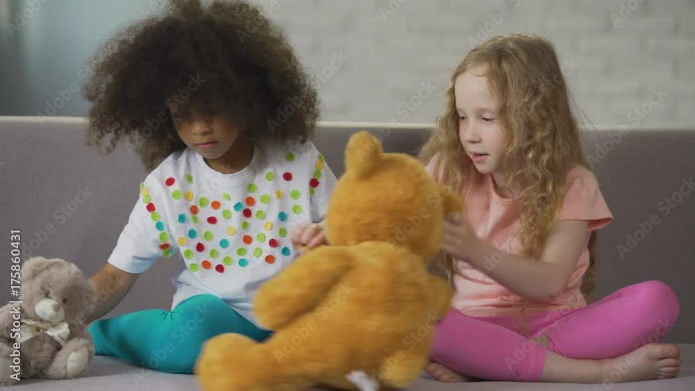 Two little multiracial girls sitting on couch and playing with teddy bears Two little multiracial girls sitting on couch and playing with teddy bears