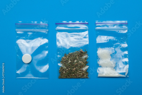 Three packets of drugs on a blue background: marijuana, cocaine and ecstasy.