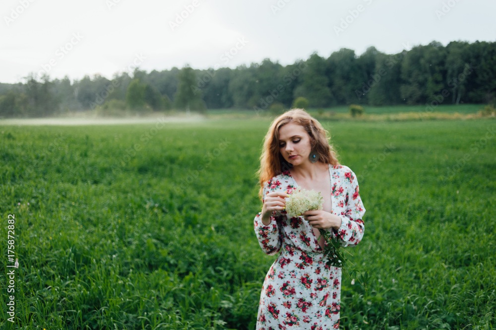 A beautiful girl in a colorful dress stands in the evening on a meadow against a background of fog.
