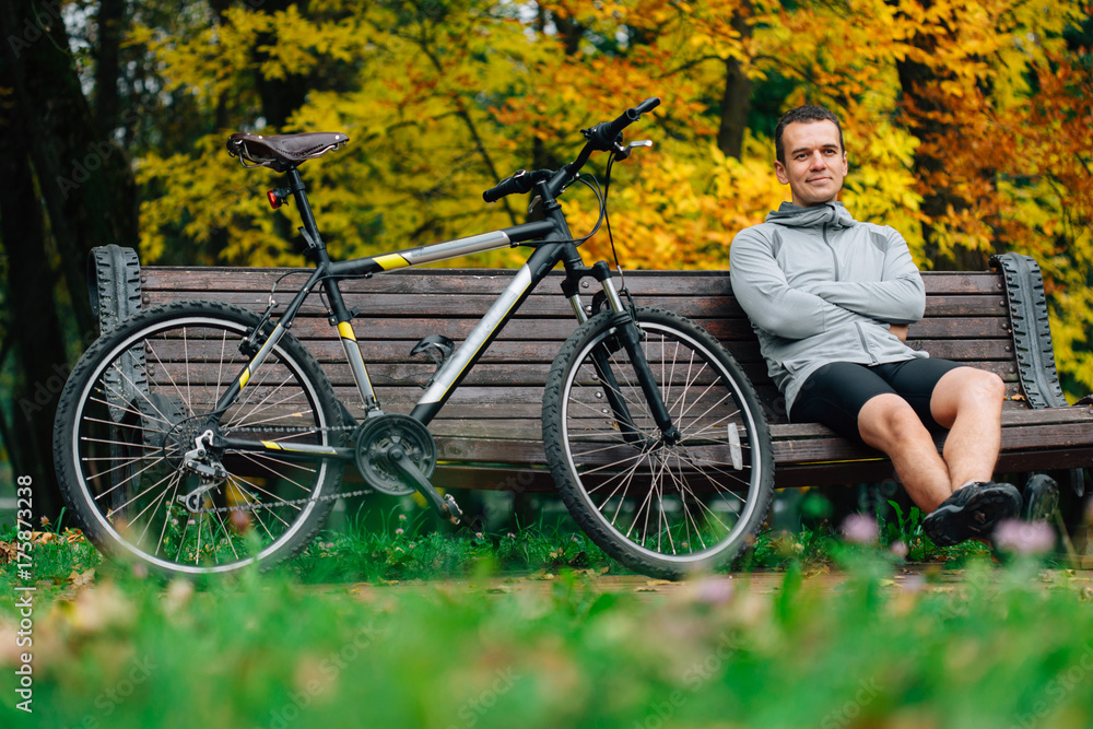 Obraz premium Young pretty athletic man sitting on bench near bicycle in colorful autumn park. Fall season background. Male cyclist resting after ride
