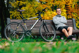 Young pretty athletic man sitting on bench near bicycle in colorful autumn park. Fall season background. Male cyclist resting after ride