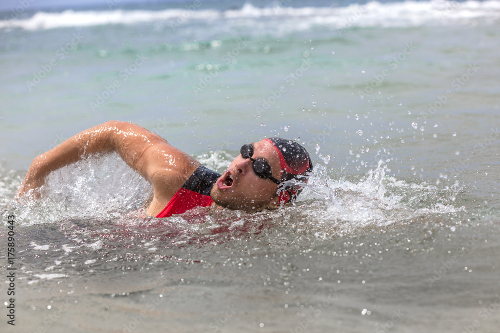 Professional triathlete man swimming crawl ocean freestyle crawl in ocean. Male triathlon swimmer wearing cap, goggles and red triathlon tri suit training for ironman breathing out of water.