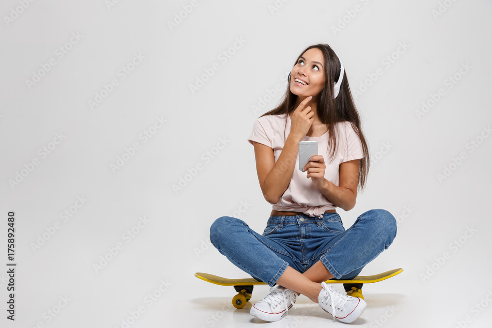Portrait of thinking young woman in white headphone holding mobile phone, sitting on skateboard, looking up Portrait of thinking young woman in white headphone holding mobile phone, sitting on skateboard, looking up