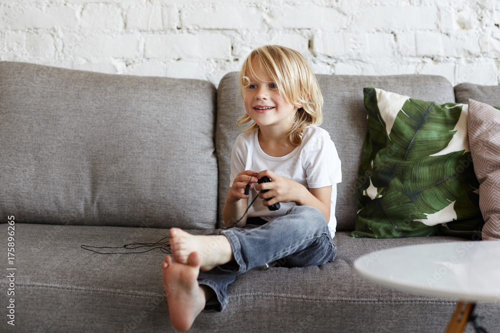 Happy adorable cute little boy with loose messy blonde hair sitting  barefooted on grey couch in living room with MP3 player or other electronic  device while relaxing at home after kindergarten Stock
