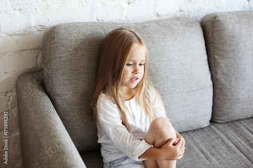 Foto High angle view of upset sweet blue eyed little girl with long straight hair sitting alone on grey couch feeling sad because of being punished by her mother for bad behaviour