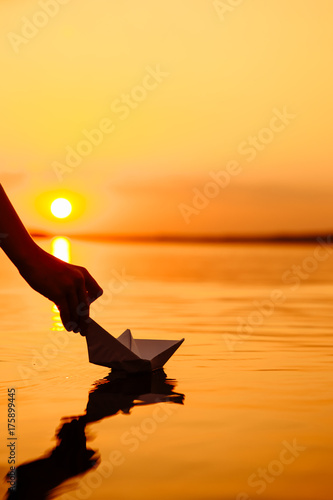 A kid putting a paper boat into water. Beautiful sunset. Origami. River. Lake.
