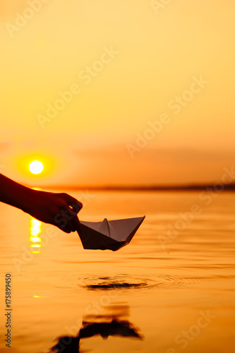 A kid putting a paper boat into water. Beautiful sunset. Origami. River. Lake.
