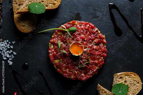 Beef tartar with a quail egg on top, grilled bread, greenery, served on a black stone plate. photo