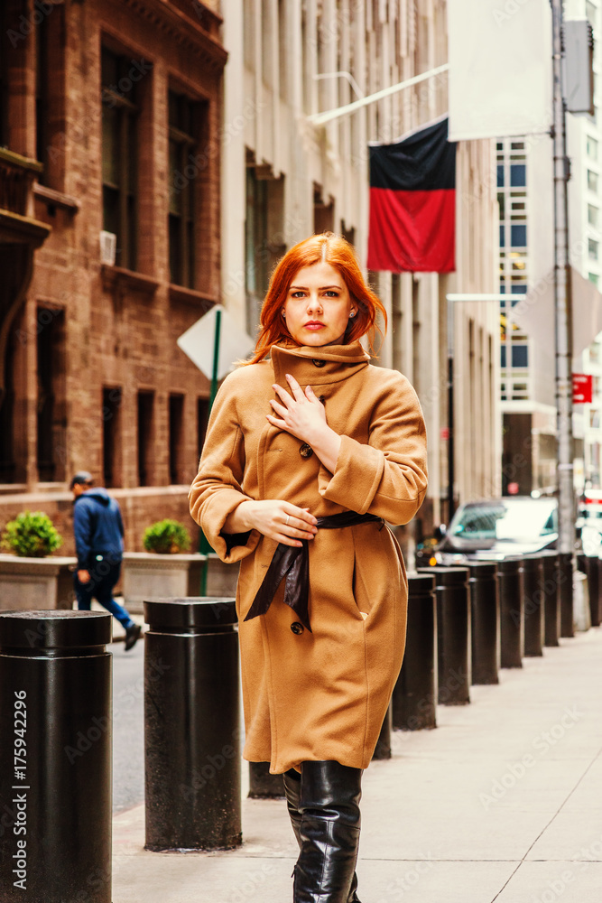 American businesswoman with red hair traveling, working in New York, wearing long brown woolen overcoat, leather boots, hand touching coat in front, walking on narrow vintage street in winter day..
