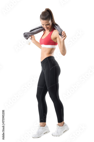 Happy fit woman in top and leggings holding towel around neck smiling and looking down. Full body length portrait isolated on white background. 