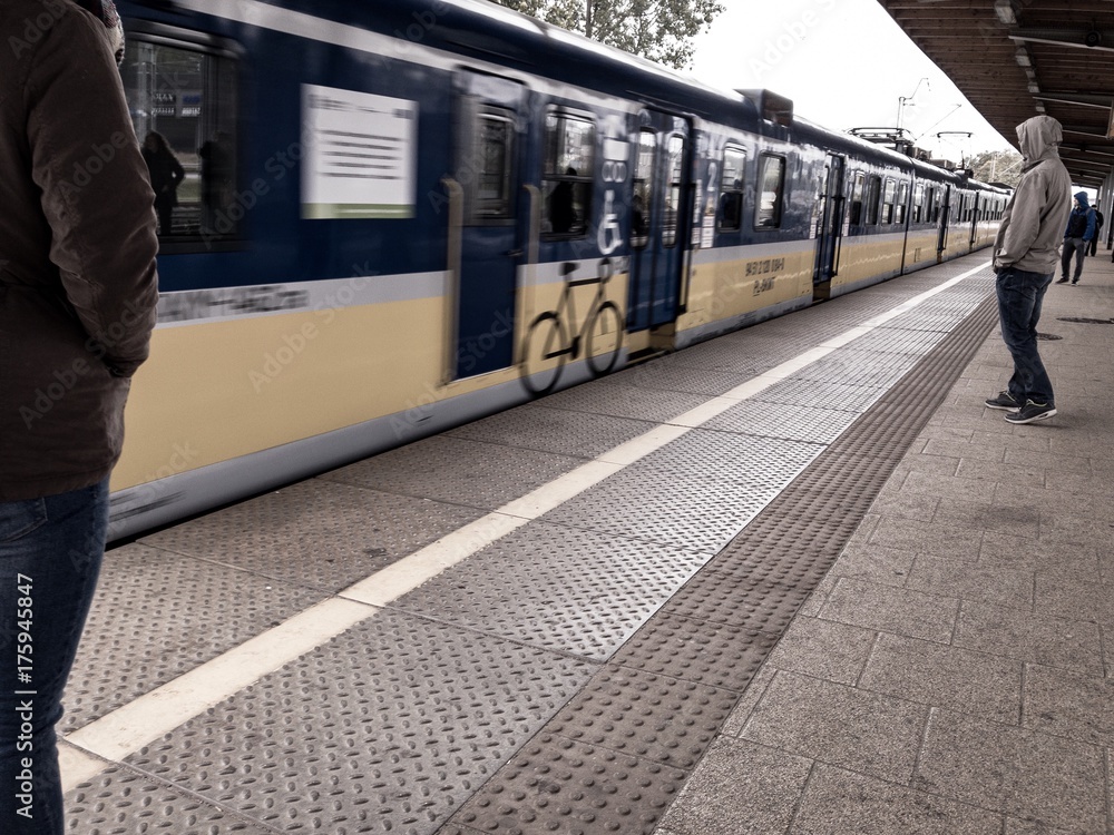 Pociąg wjeżdża na stacje kolejową w dzień, na peronie stoi mężczyzna w bluzie z kapturem zaciągniętym na głowę, pociąg jest w ruchu 