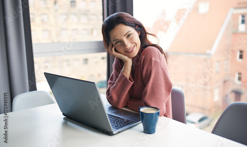 Young attractive girl sitting near big window with coffee and working on laptop.