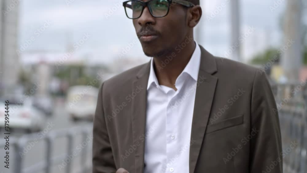 Tilt up of African businessman in eyeglasses and formal suit standing on the side of street and typing on smartphone Tilt up of African businessman in eyeglasses and formal suit standing on the side of street and typing on smartphone