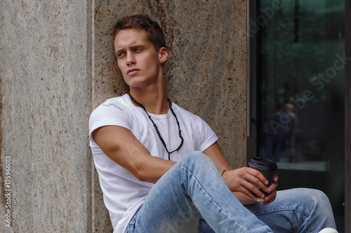 A young white man in a white T-shirt and blue jeans sits leaning against a wall, holding a paper glass with coffee in his hands.