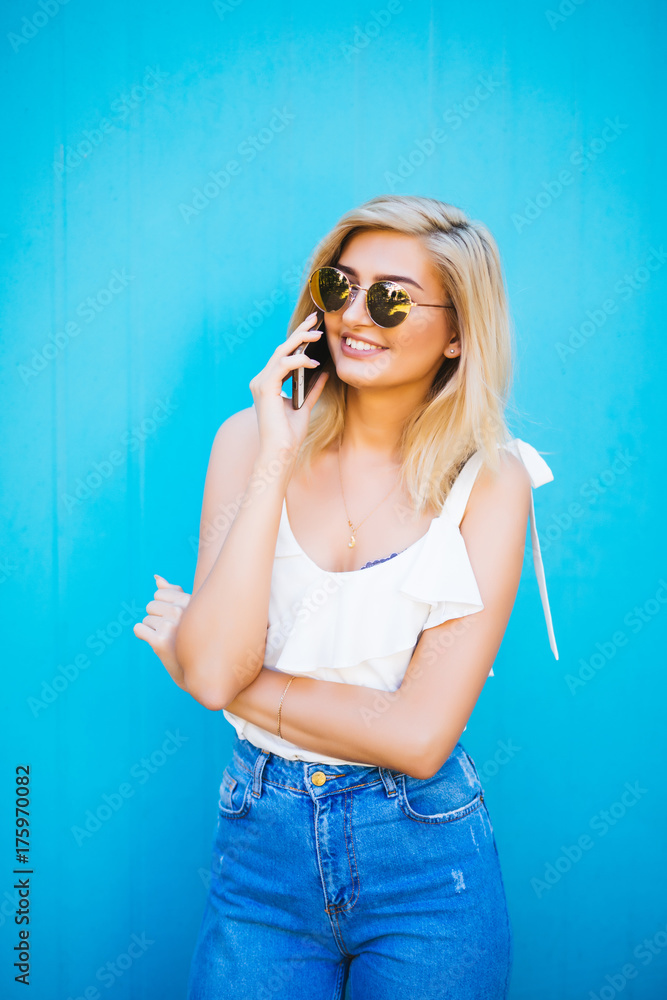 Fototapeta premium Pretty Woman Calling Someone Through Mobile Phone While Smiling at the Camera Against Blue Wall Background with Copy Space.
