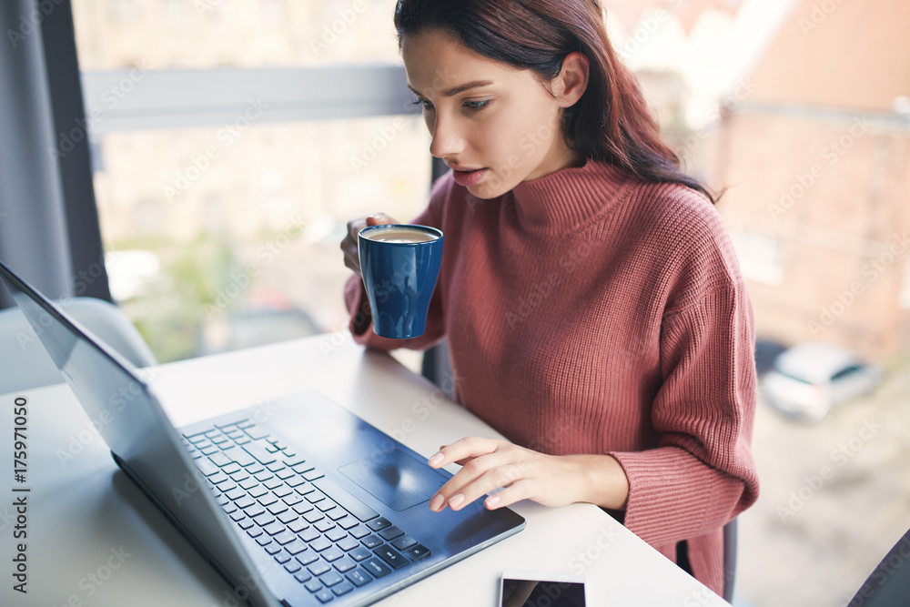 Fototapeta premium Young attractive girl sitting near big window with coffee and working on laptop.