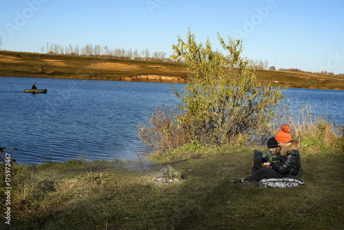 boy and girl sitting on the shore of a pond with a laptop in hands autumn day