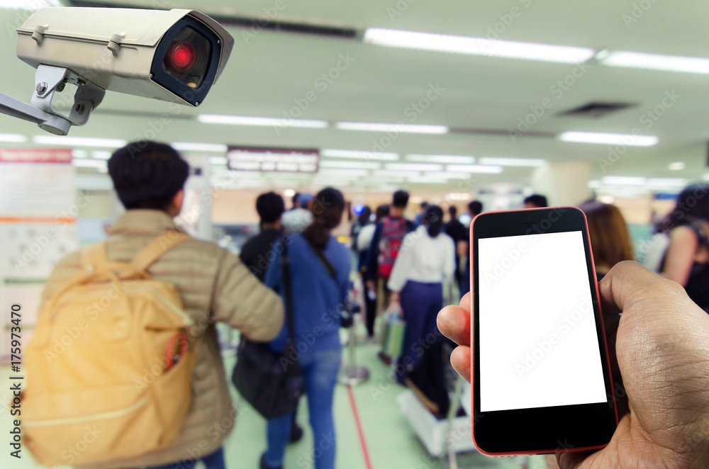 hand holding mobile smart phone and CCTV security indoor camera system operating with blurred image of people queue at immigration control at airport,