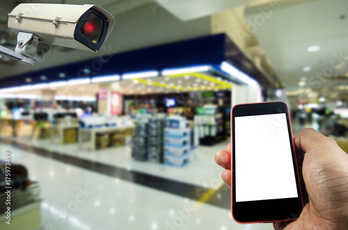 hand holding mobile smart phone and CCTV security indoor camera system operating with blurred image in supermarket or shopping mall, internet, surveillance security, safety technology concept