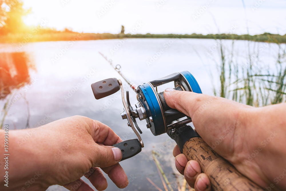 A man is fishing with a backcasting reel. Hands, a rod and a backcasting reel in the background of the rising sun