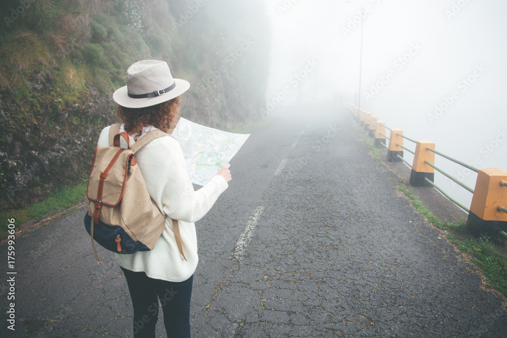 Young curly travelling woman wearing backpack and hat standing on the road in fog, searching trail and using map.