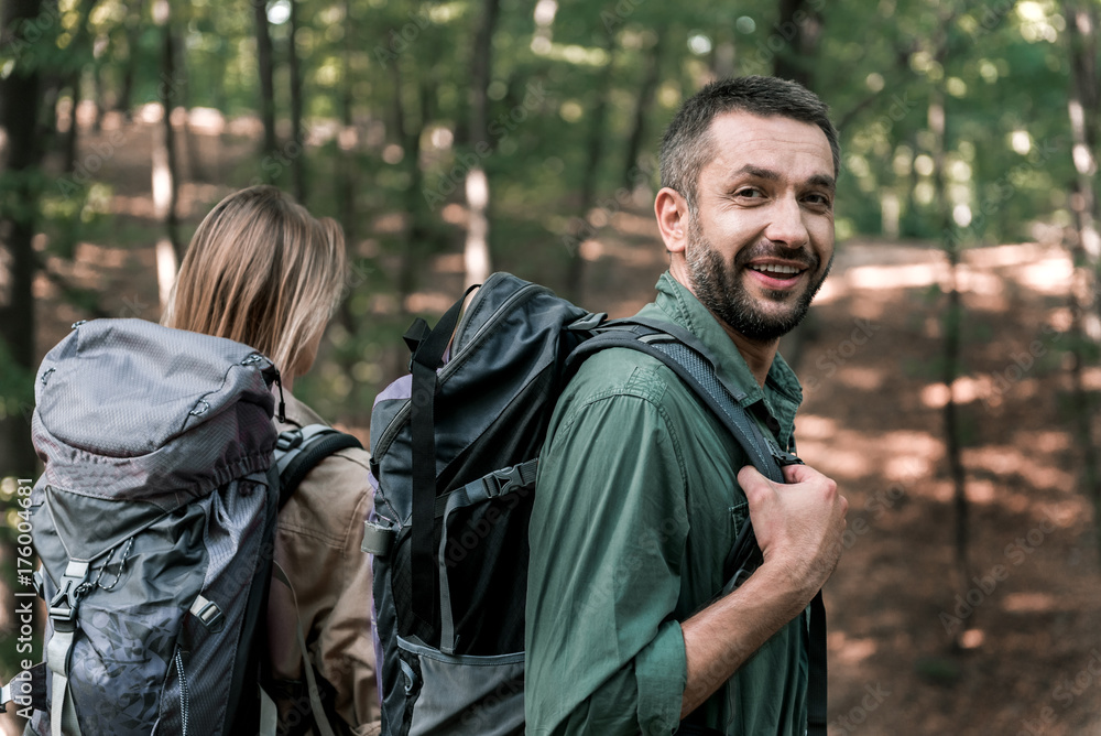 Cheerful couple hiking in forest with rucksacks