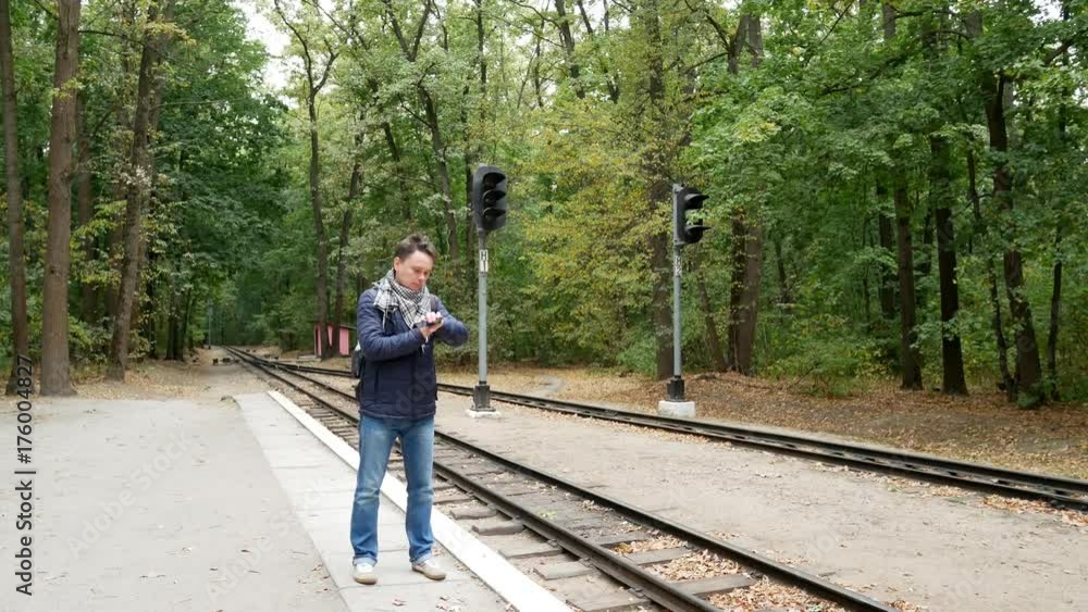 A young man is waiting for a train on an apron and looks at a wristwatch. A railroad station in the forest in the autumn. Student traveler is nervous because of late train A young man is waiting for a train on an apron and looks at a wristwatch. A railroad station in the forest in the autumn. Student traveler is nervous because of late train