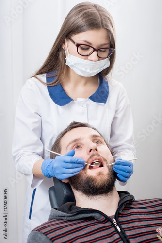 Young female dentist holding dental tools - mirror and probe and doing first check-up male patient in dental office. Doctor wearing white uniform, glasses, mask and blue gloves. Dentistry