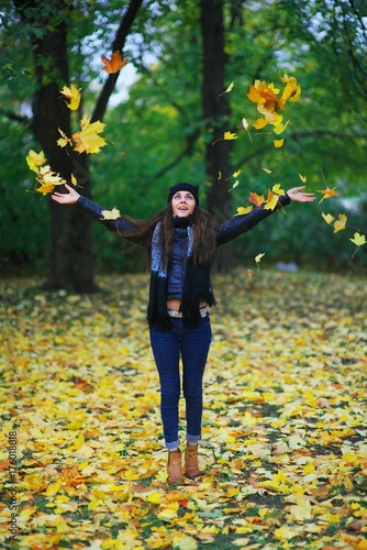A girl in a hat, jacket, jeans and a scarf throws yellow autumn leaves. She is cheerful and joyful