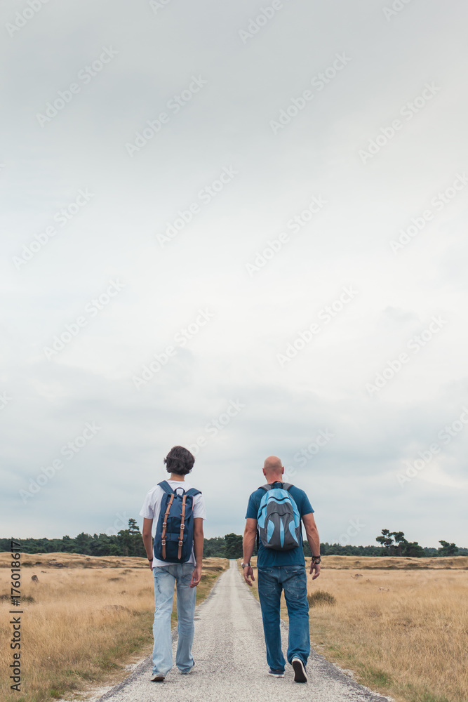 Two men hiking with backpacks on a long road in the fields Two men hiking with backpacks on a long road in the fields