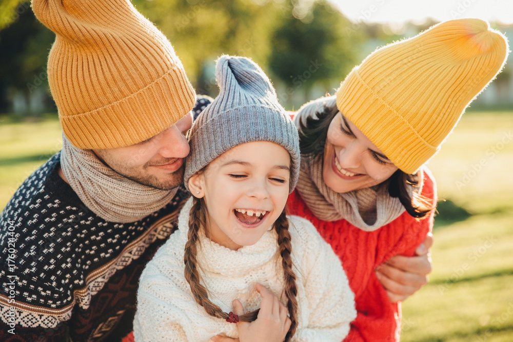 Obraz premium Adorable funny small kid has fun with parents who look at her with great love, enjoy spending free time together, admire beautiful nature and fresh air. People, happiness and childhood concept